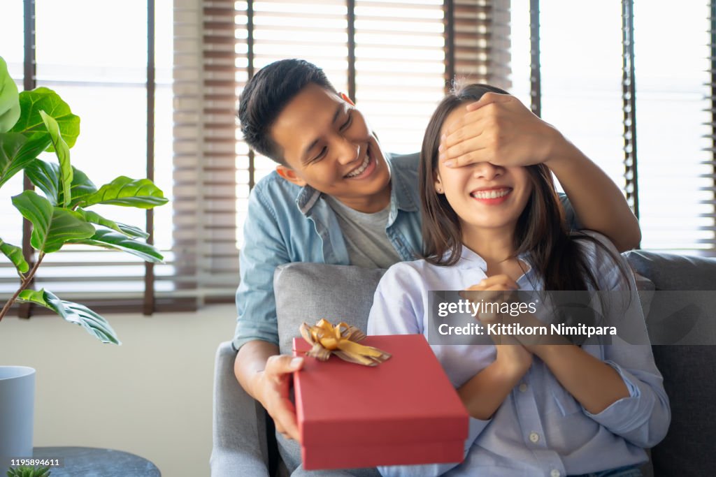 Image of young guy with giftbox closing his girlfriend eyes to make a surprise for her.