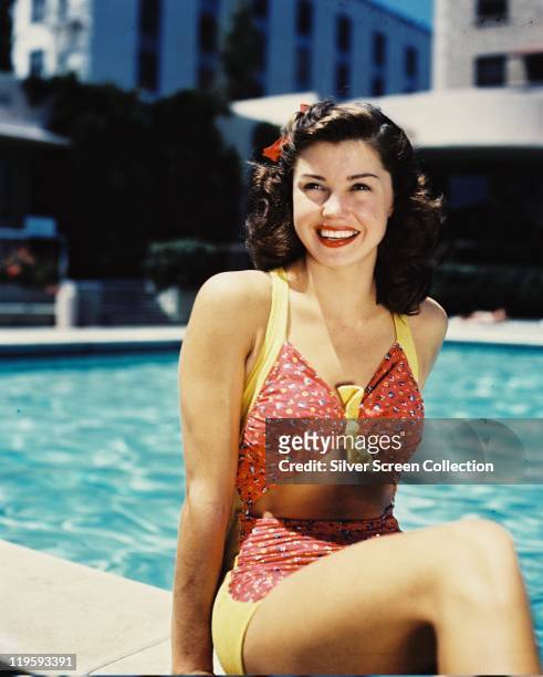 Esther Williams, US actress and former Olympic swimmer, wearing a red-and-yellow bathing costume, smiling as she poses at the edge of a swmiming...
