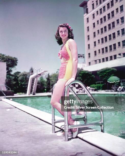 Esther Williams, US actress and former Olympic swimmer, wearing a bathing costume as she poses leaning against the ladder at the edge of a swmiming...
