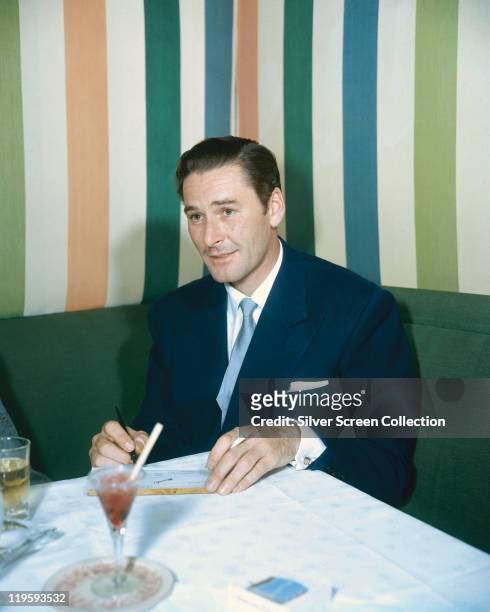Errol Flynn , Australian actor, sitting on a green seat and sitting at a table, on which drinks rest, with striped wallpaper in the background, 1950.