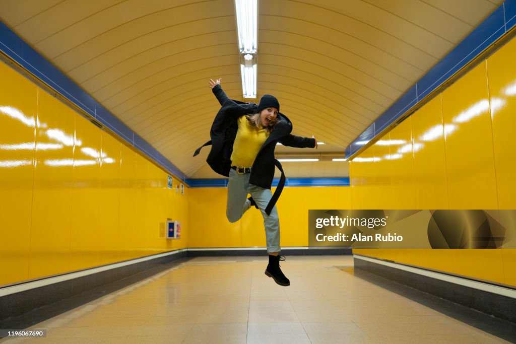 Young blonde girl in the subway with hat and black clothes waiting for the subway with yellow background jumping