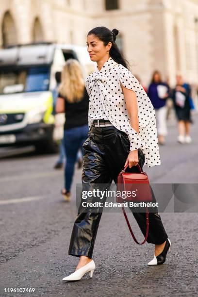 Caroline Issa wears a white shirt with printed black polka dots, black shiny vinyl pants, a red leather bag, black and white pointy shoes, during...