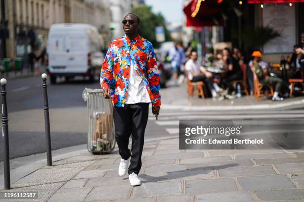 Guest wears sunglasses, a white t-shirt, a colorful floral print shirt, black pants, white sneakers, outside Sankuanz, during Paris Fashion Week -...