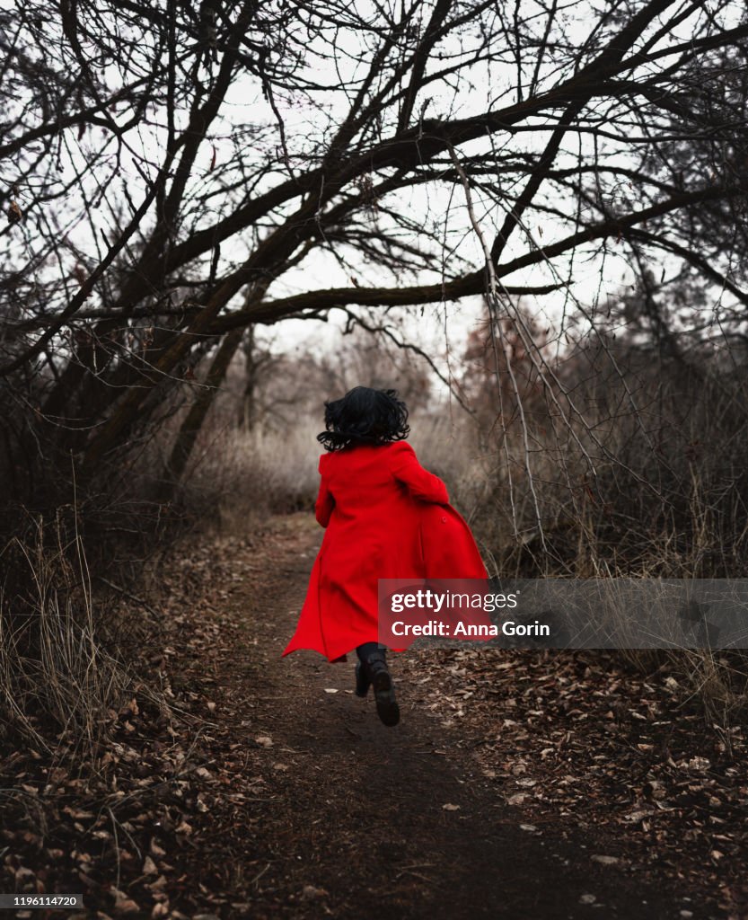 Back view of woman with medium-length black hair in long red coat running down forest path in winter, desaturated effect