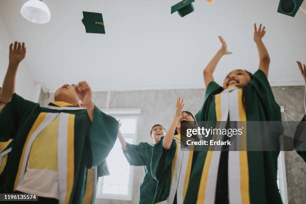 grand groupe d'étudiants heureux d'élémentaire célébrant leur jour de graduation à l'intérieur tout en jetant leurs chapeaux vers le haut en l'air. - the asian awards photos et images de collection