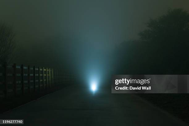 a mysterious spooky glowing light on a road, with a shallow depth of field. on a moody, foggy, atmospheric winters night. - road light trail stock pictures, royalty-free photos & images