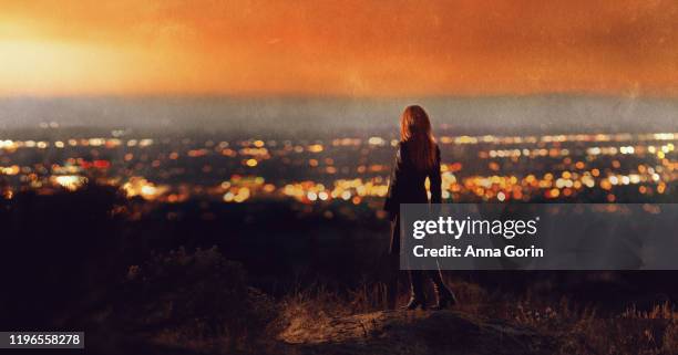 back view of woman with long red hair wearing black coat and heeled boots standing on hillside looking at sunset over valley of city lights - back of womens heads stock-fotos und bilder