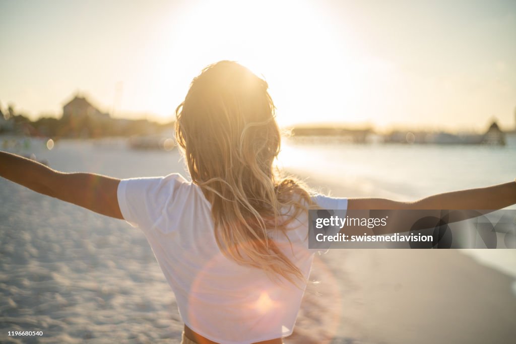 Fröhliche junge Frau umarmt die Natur bei Sonnenuntergang; Hündin steht auf Strandarmen ausgestreckt