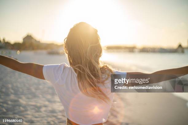 cheerful young woman embracing nature at sunset; female standing on beach arms outstretched - spirituality stock pictures, royalty-free photos & images