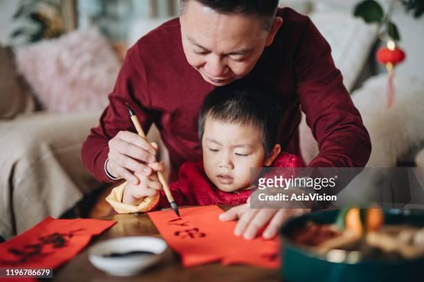 großvater übt chinesische kalligraphie für chinesisches neujahr fai chun (ausserliche botschaften) und lehrt seinen enkel, indem er es auf ein stück rotes papier schreibt - tradition stock-fotos und bilder