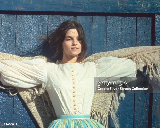 Genevieve Bujold, Canadian actress, wearing a white blouse with her arms outstretched either side of her, with a shawl, standing against a blue...