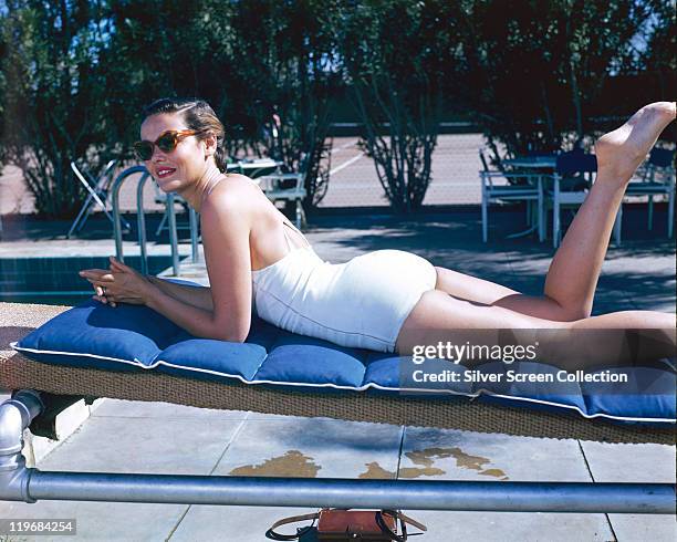 Gene Tierney , US actress, laying on her front, sunbathing on a sunlounger, wearing a white swimsuit and sunglasses, circa 1940.