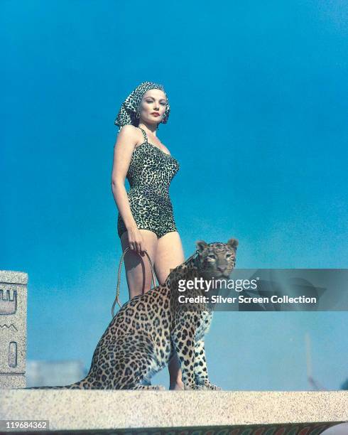 Gene Tierney , US actress, wearing a leopard print swimsuit and headscarf, posing alongside a leopard which Tierney holds on a lead, 27th July 1954.