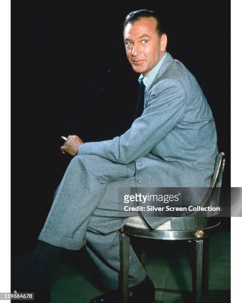 Gary Cooper , US actor, smoking a cigarette while sitting in a chair, wearing a light grey suit, in a studio portrait, circa 1955.