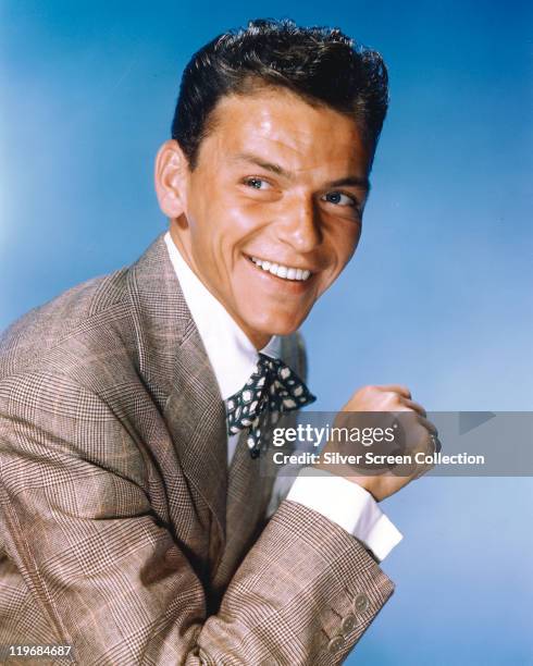 Frank Sinatra , US singer and actor, smiling while dressed in a grey jacket, a white shirt and a blue-and-white bow tie in a studio portrait, against...