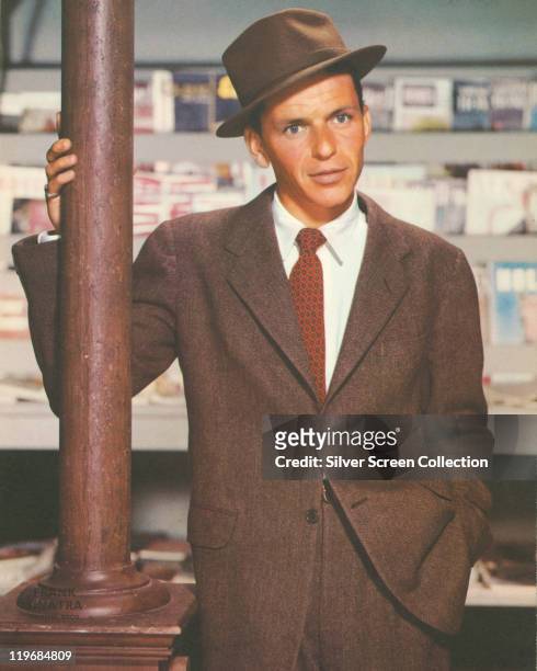 Frank Sinatra , US singer and actor, dressed in a brown jacket, brown fedora, white shirt and a red-and-black tie, leaning against a lamppost, circa...