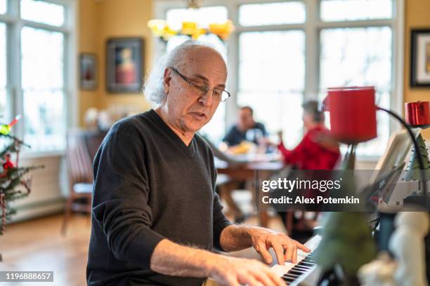 senior man playing piano when his family dinning around a table in the backdrop - keyboard instrument stock pictures, royalty-free photos & images