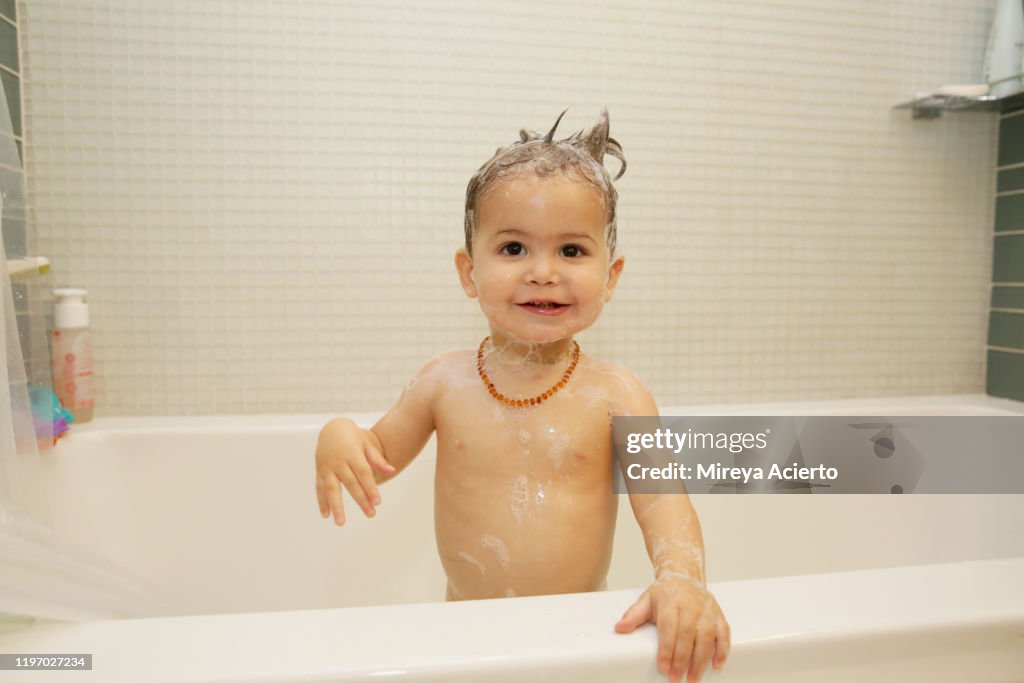 A LatinX toddler stands in the bathtub smiling, wearing a beaded necklace and a head full of soap.