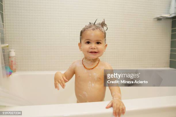 A LatinX toddler stands in the bathtub smiling, wearing a beaded necklace and a head full of soap.