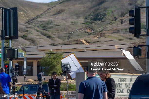 Journalists report from a parking lot of the Church In the Canyon as investigators on a distant hill work at the scene of the helicopter crash, where...
