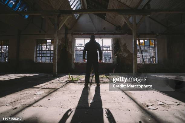 uomo adulto in piedi all'interno di un grande edificio buio, spettrale e abbandonato illuminato dalla luce del sole attraverso la finestra - omicidi seriali foto e immagini stock