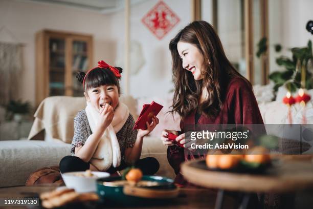 lovely daughter enjoying traditional snacks while helping her mother to prepare red envelops (lai see) at home for chinese new year - chinese new year stock pictures, royalty-free photos & images