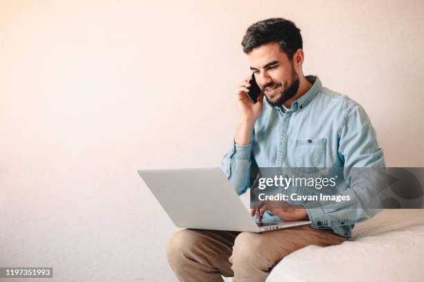 happy businessman talking on smart phone while using laptop at home - em frente de imagens e fotografias de stock