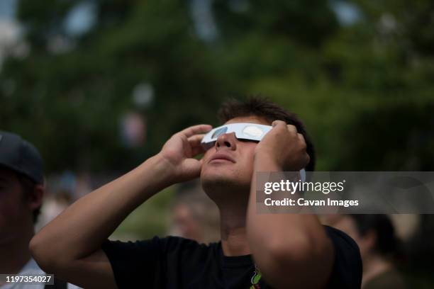 young man looking up at solar eclipse wearing paper protective glasses - solar eclipse stock-fotos und bilder