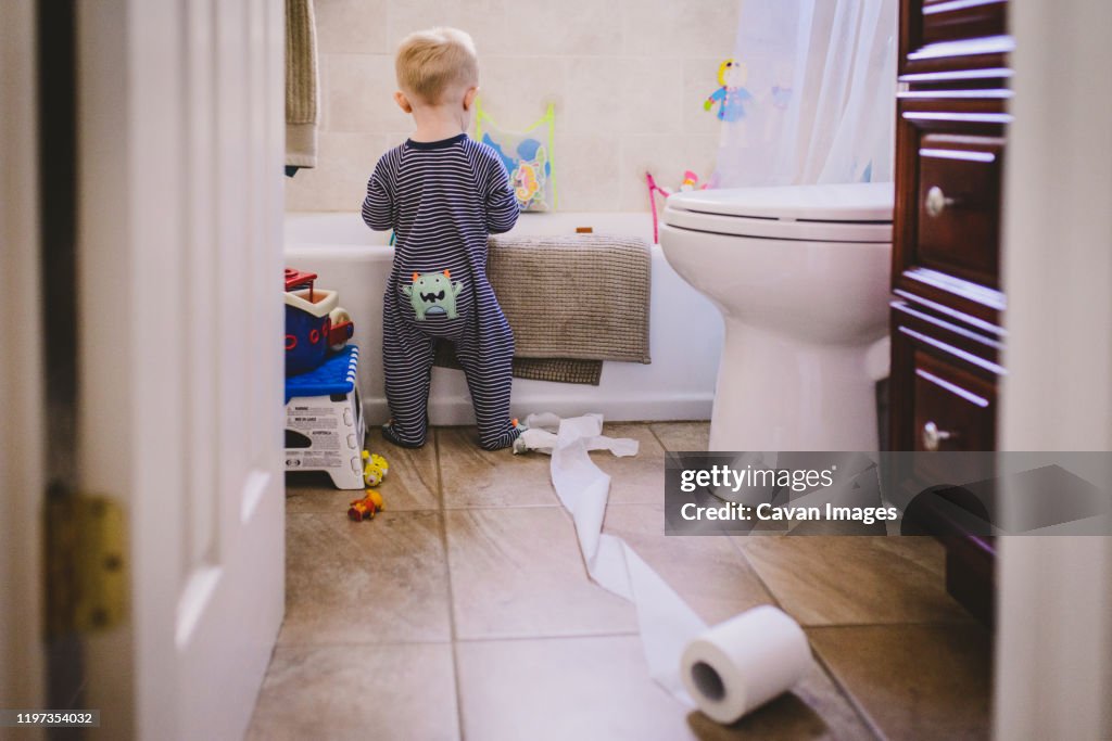 A baby boy stands in a bathroom with an unfurled toilet roll.