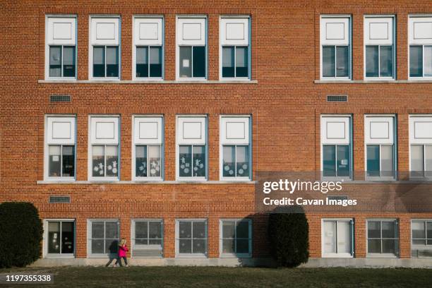 a little girl in a pink coat walks past a school building. - public school building stock pictures, royalty-free photos & images