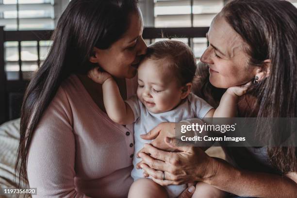 happy baby girl holding cheeks of both smiling moms in front of window - diverse family stock-fotos und bilder