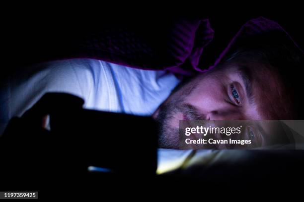 bearded young man is lying in bed under his blanket looking at phone - homens de idade mediana imagens e fotografias de stock