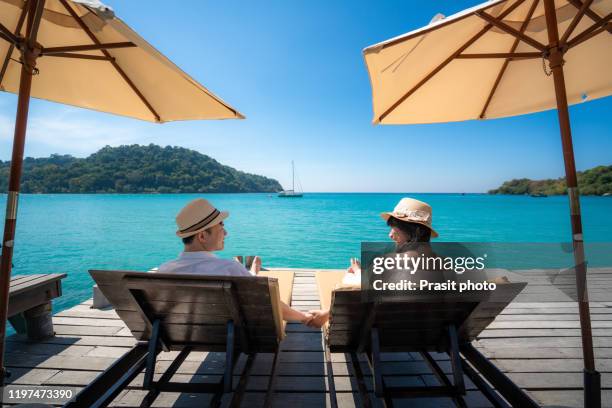 asian happy couple relaxing on chair beach with umbralla on wooden deck over sea background during summer vacation together in koh kood, trat, thailand. - man on the beach relaxing in deckchair fotografías e imágenes de stock