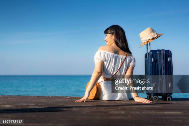 asian happy woman tourist sitting on wooden deck on the summer beach with luggage bag and looks into the distance to the sea. summer travel vacation and holidays concept. - happy holidays around the world stock pictures, royalty-free photos & images