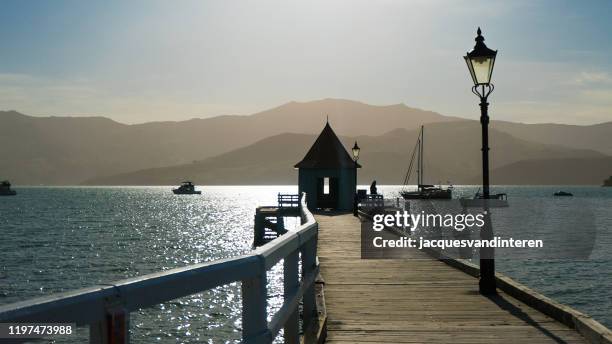 hafenanleger mit kiosk und laternen. boote auf dem wasser und berge im hintergrund. dämmerung und hintergrundbeleuchtung - akaroa stock-fotos und bilder