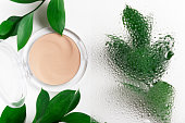 Cosmetic container and plant twigs top view composition. Makeup attribute, natural cosmetology product concept. Leafage behind wet glass on white background. Face powder, foundation, concealer jar