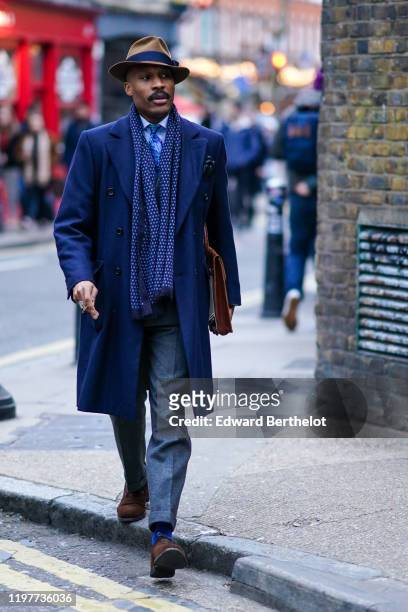 Guest wears a brown hat, a scarf, a blue long coat, a shirt, a tie, gray pants, brown shoes, a brown clutch, during London Fashion Week Men's January...