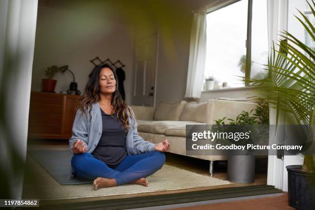 mature woman meditating while practicing yoga in her living room - mature woman yoga indoors stock pictures, royalty-free photos & images