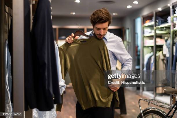 un tipo latinoamericano probando un suéter encima en una tienda de ropa para hombres - ropa fotografías e imágenes de stock