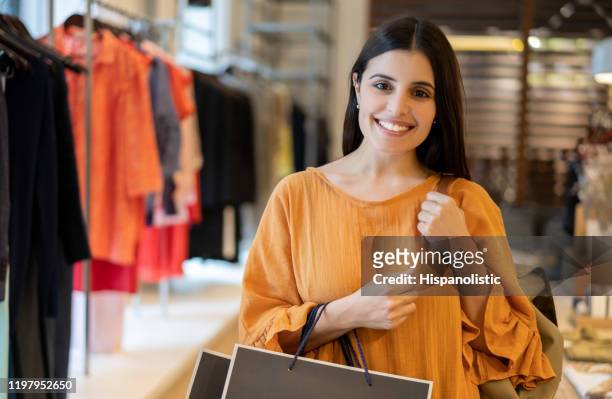 vacker latinamerikansk kvinna på en klädbutik som håller shoppingväskor leende på kamera - latin american and hispanic shopping bags bildbanksfoton och bilder