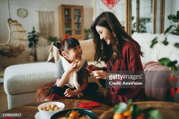 lovely daughter enjoying traditional snacks while helping her mother to prepare red envelops (lai see) at home for chinese new year - chinese prepare for lunar new year stock pictures, royalty-free photos & images