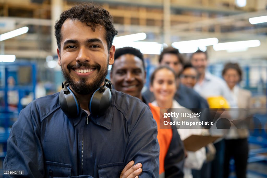 Vrolijke knappe blauwe kraag werker en team van ingenieurs in een fabriek staand in een rij glimlachend op camera