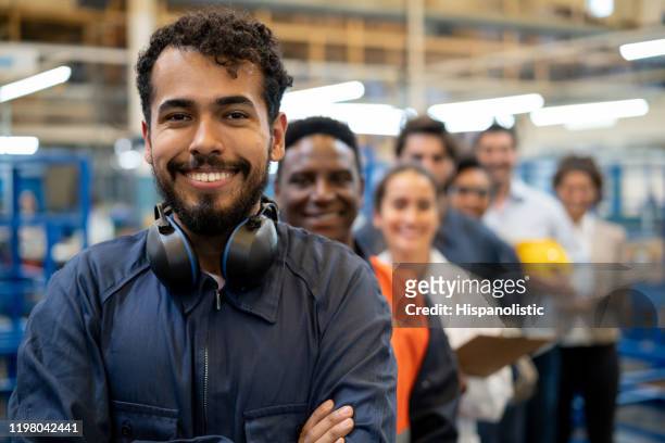 cheerful handsome blue collar worker and team of engineers at a factory standing in a row smiling at camera - manufacturing stock pictures, royalty-free photos & images