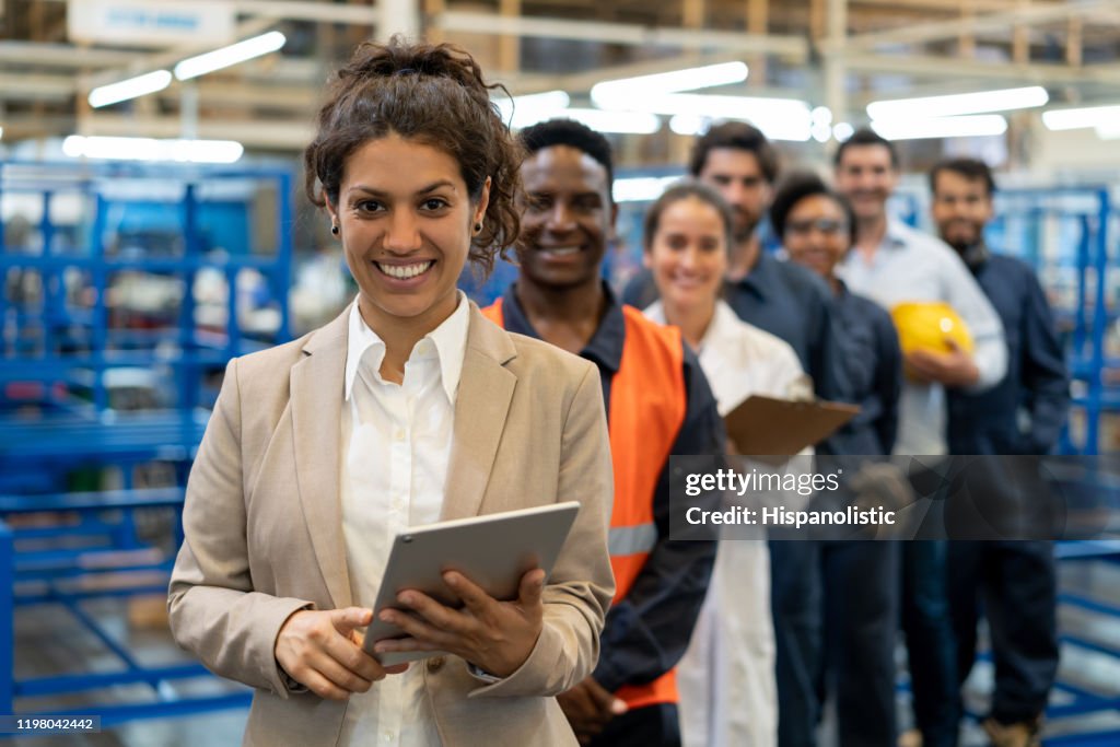 Schöne weibliche Managerin in einer Fabrik, die ein Tablet und ein Team von Arbeitern, Ingenieuren und Inspektoren hält, die in einer Reihe stehen und vor der Kamera lächeln