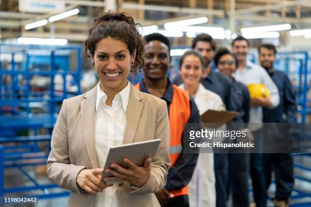 schöne weibliche managerin in einer fabrik, die ein tablet und ein team von arbeitern, ingenieuren und inspektoren hält, die in einer reihe stehen und vor der kamera lächeln - berufliche beschäftigung stock-fotos und bilder