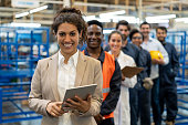 Beautiful female manager at a factory holding a tablet and team of blue collar workers, engineers and inspectors standing in a row smiling at camera