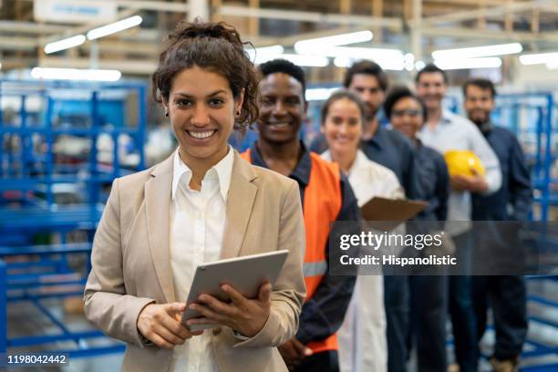 beautiful female manager at a factory holding a tablet and team of blue collar workers, engineers and inspectors standing in a row smiling at camera - engineering team stock pictures, royalty-free photos & images