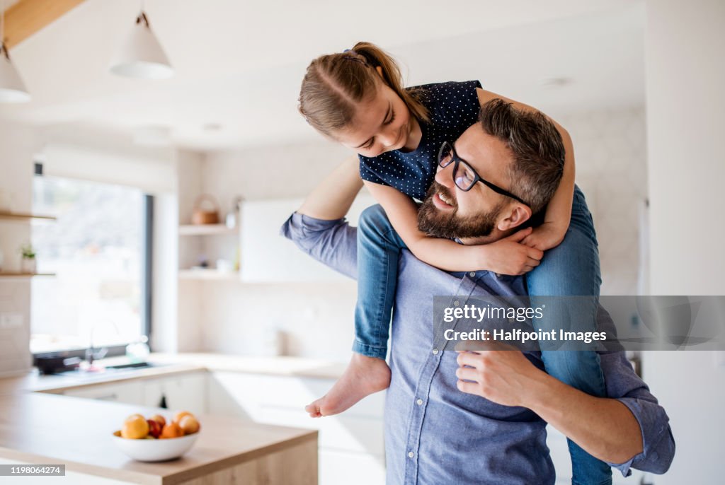 Mature father giving piggyback ride to small daughter indoors at home.