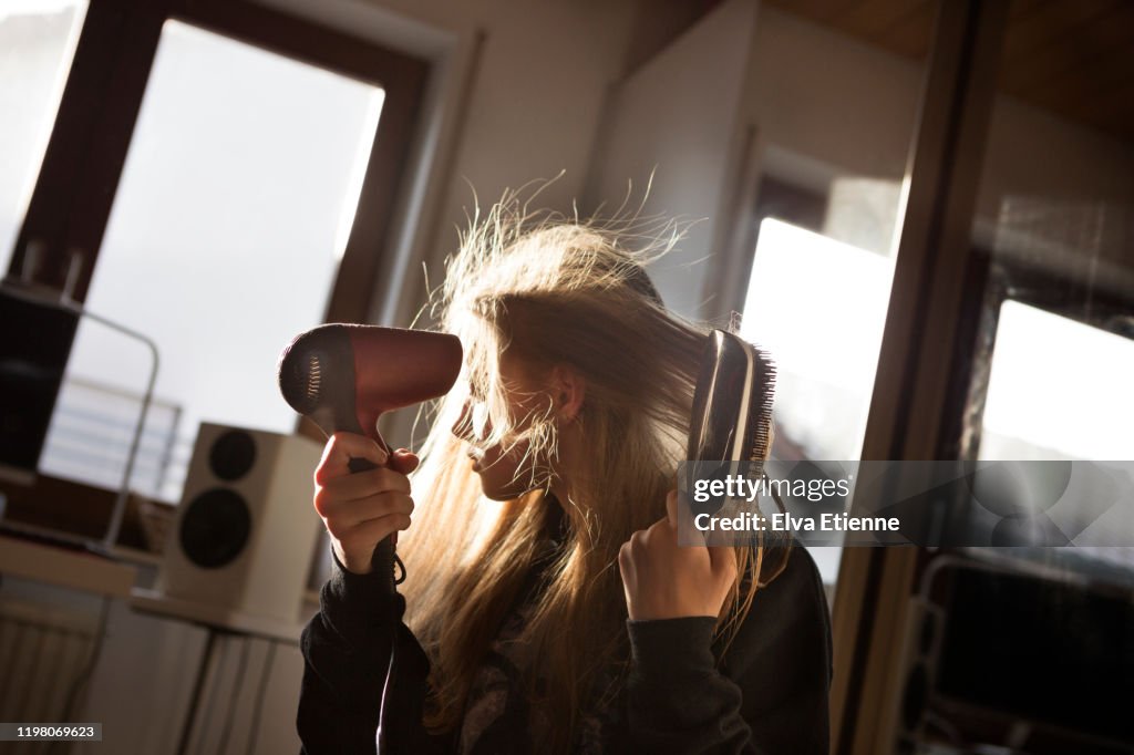 Girl (12-13) blow drying her long hair with an electric hairdryer in a bedroom