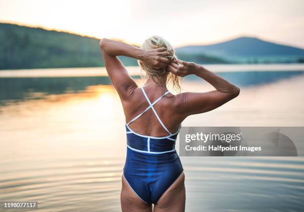 rear view of active senior woman standing by water outdoors in nature at sunset. - old woman in swimsuit stock pictures, royalty-free photos & images